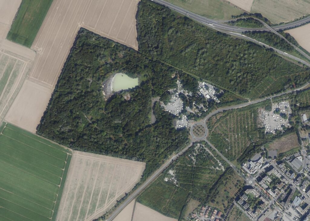 Aerial view of the former Mirapolis site at Courdimanche today, showing dense woodland covering the old park, a small lake, clusters of caravans and mobile homes, farm fields to the west and the edge of Cergy-Pontoise to the south-east.