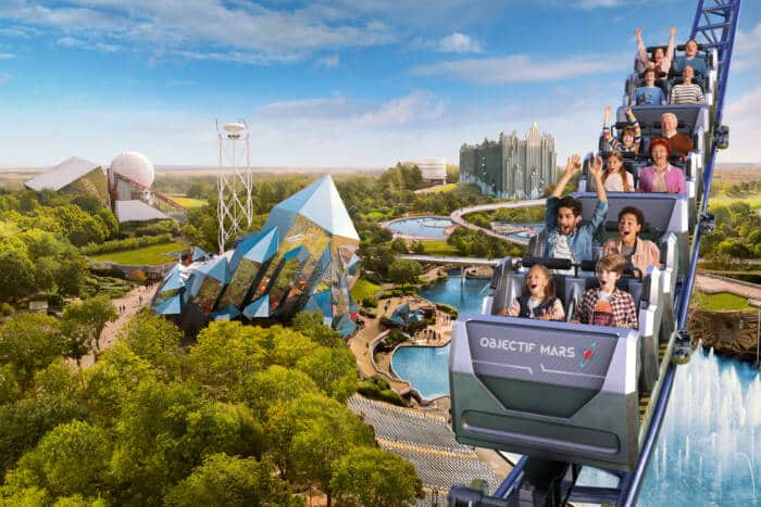Futuroscope's Objectif Mars roller coaster in action, with riders cheering as the train banks past the park's geometric pavilions and lake.