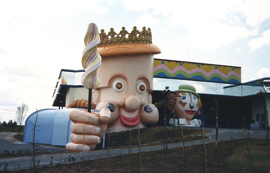 A giant sculpture of Dame Tartine at the Mirapolis theme park in 1987: a huge brioche-shaped head wearing a gold crown, holding an oversized striped candy cane, in front of a park building.