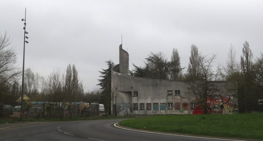 The derelict concrete entrance building of the abandoned Mirapolis theme park at Courdimanche in 2025, its windows broken and walls covered in graffiti, with a curving road and bare trees in front of it.