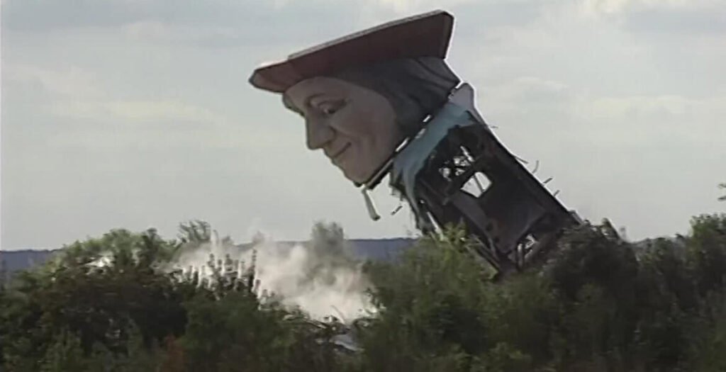 The thirty-five-metre Gargantua statue from Mirapolis tipping sideways as it collapses in 1995, its wide-brimmed hat and painted face still intact, dust rising from the trees below.