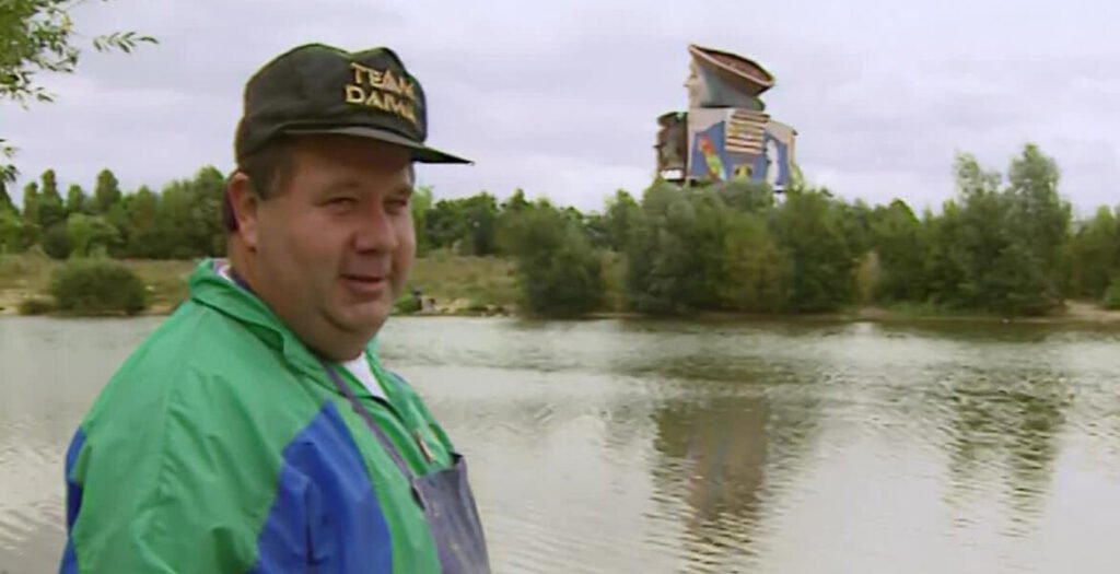 A fisherman in a green and blue jacket beside a lake at Courdimanche in 1995, with the thirty-five-metre Gargantua statue from the closed Mirapolis theme park visible above the treeline behind him.
