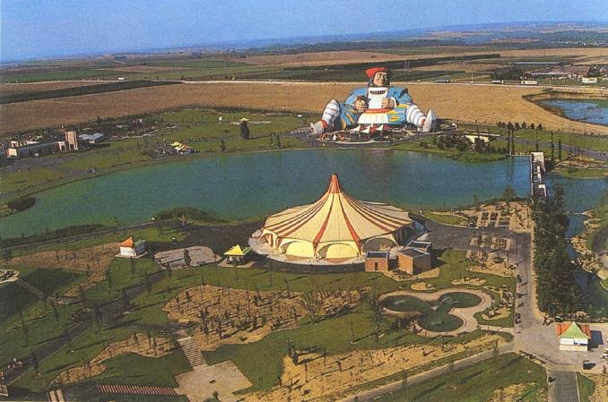 Aerial photograph of the Mirapolis theme park in 1987, showing a red and white big top tent beside a lake, with the thirty-five-metre Gargantua statue standing at the far edge of the site.